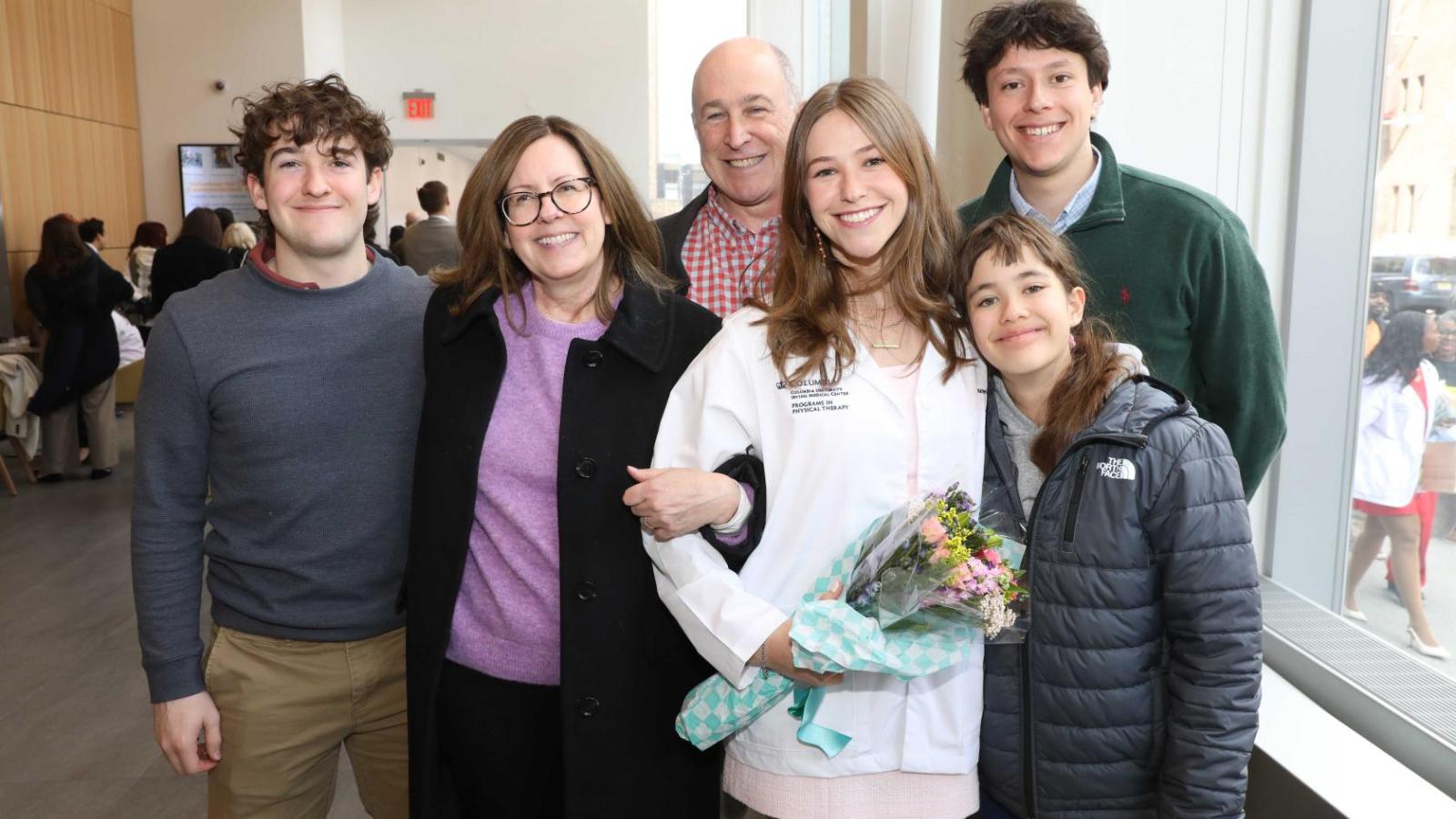 Columbia physical therapy student in a white coat poses with family members while holding flowers after the ceremony.