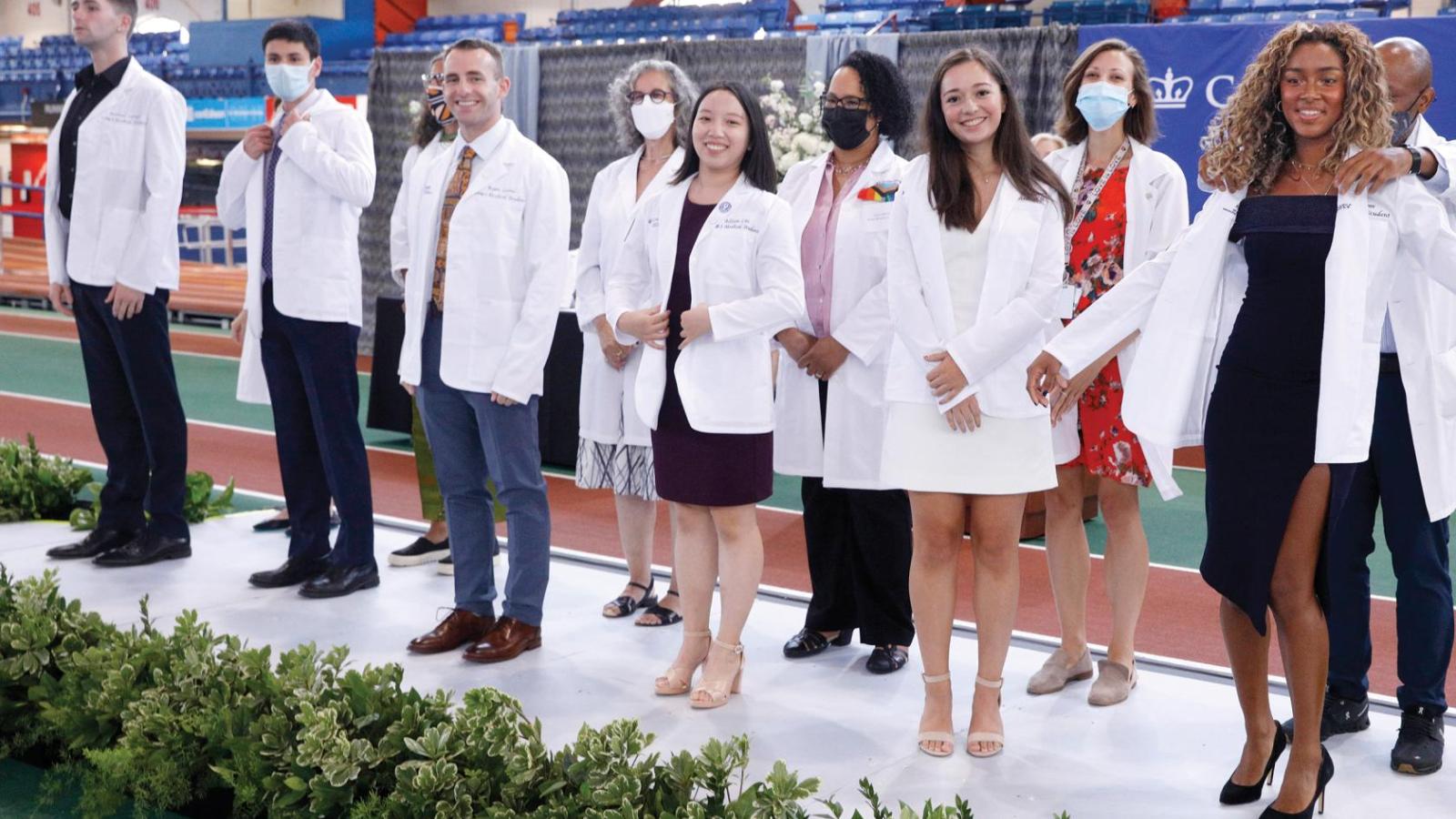 Students wear white coats and stand on a stage