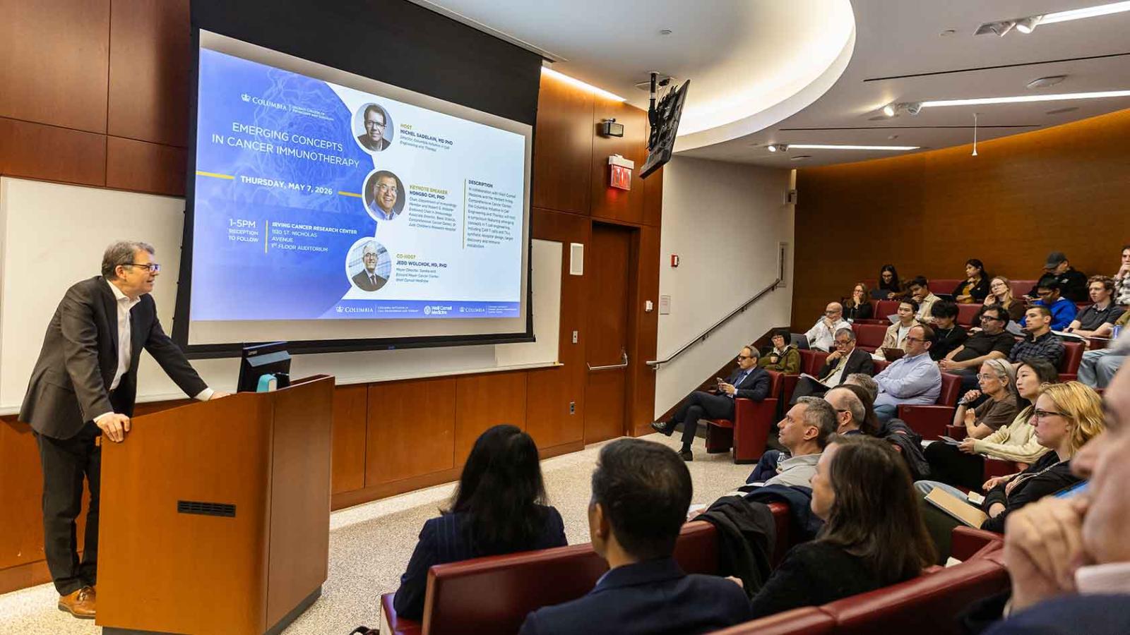 A speaker stands at a podium in a packed auditorium presenting a cancer immunotherapy symposium.