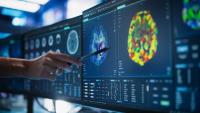 A researcher points to colorful brain imaging scans displayed across multiple monitors in a lab.