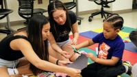 OT students help a child cut paper with scissors.