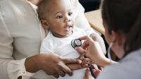 Toddler sits on parent's lap while pediatrician listens with stethoscope.