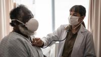 Doctor examines elderly African American man with stethoscope, both wearing masks.