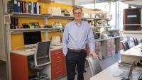 Man standing in a laboratory office with shelves and equipment behind him.