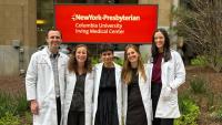 Group of five clinicians in white lab coats standing together and smiling outside in front of a NewYork-Presbyterian Columbia University Irving Medical Center sign.